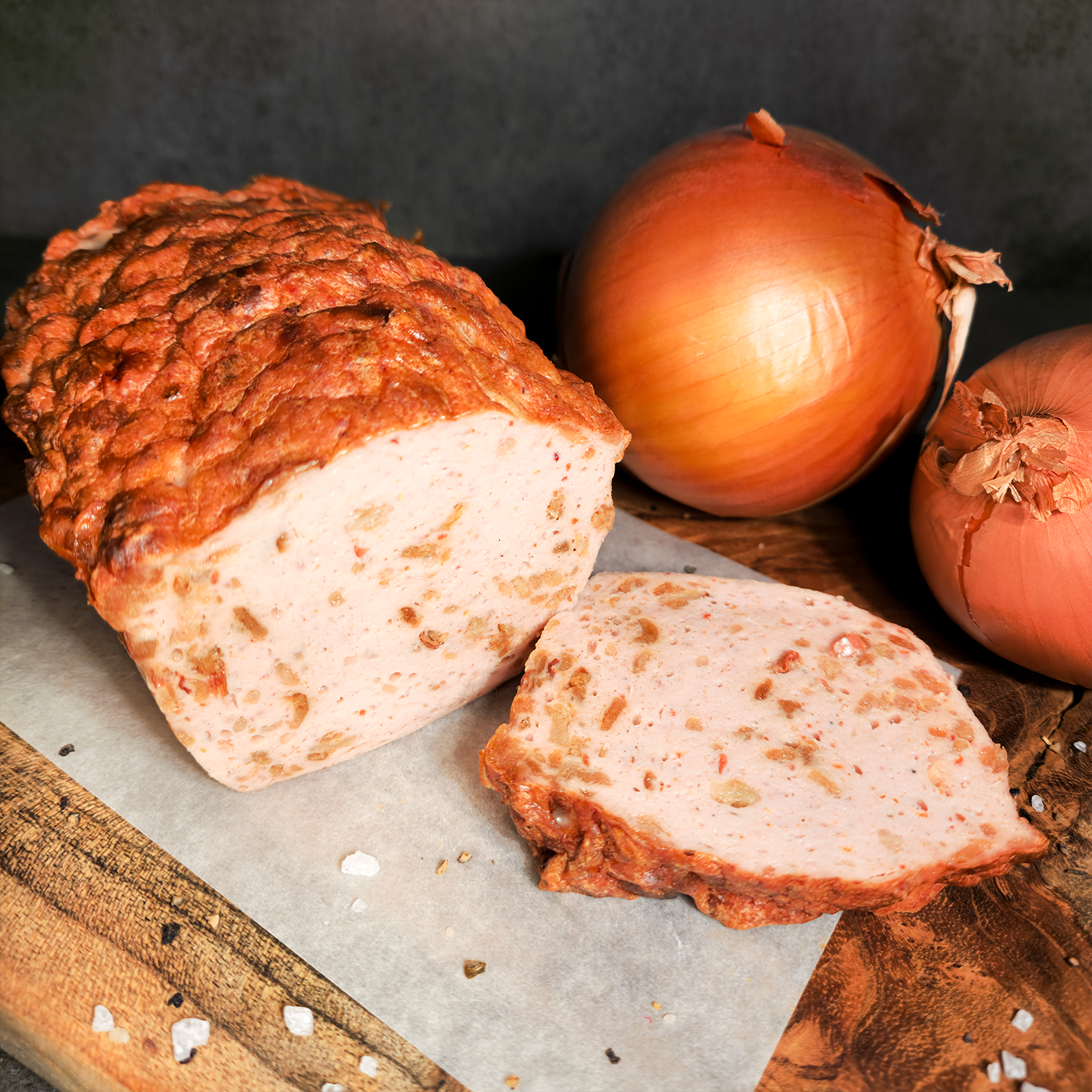 Würziger Zwiebelwiegebraten online bestellen. Zwiebelwiegebraten aufgeschnitten auf Holzbrett, im Hintergrund eine Zwiebel als Deko.