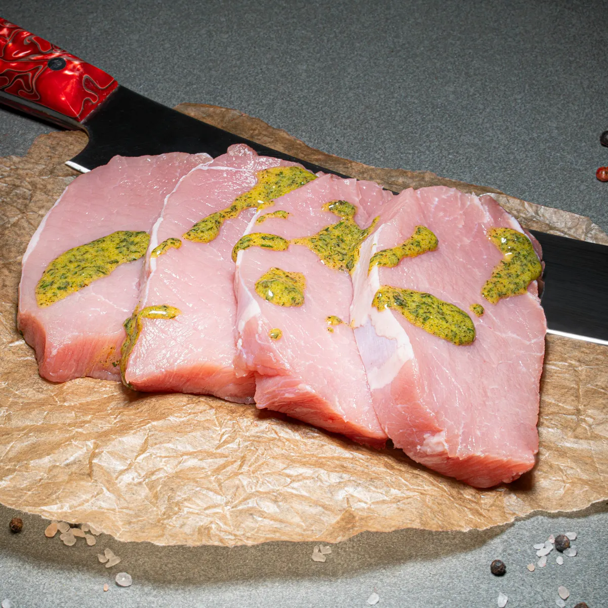 Schweinerückensteaks mit Kräutermarinade fuer den Grill, auf Fleischpapier, im Hintergrund ist ein FLeischmesser mit rotem Griff.