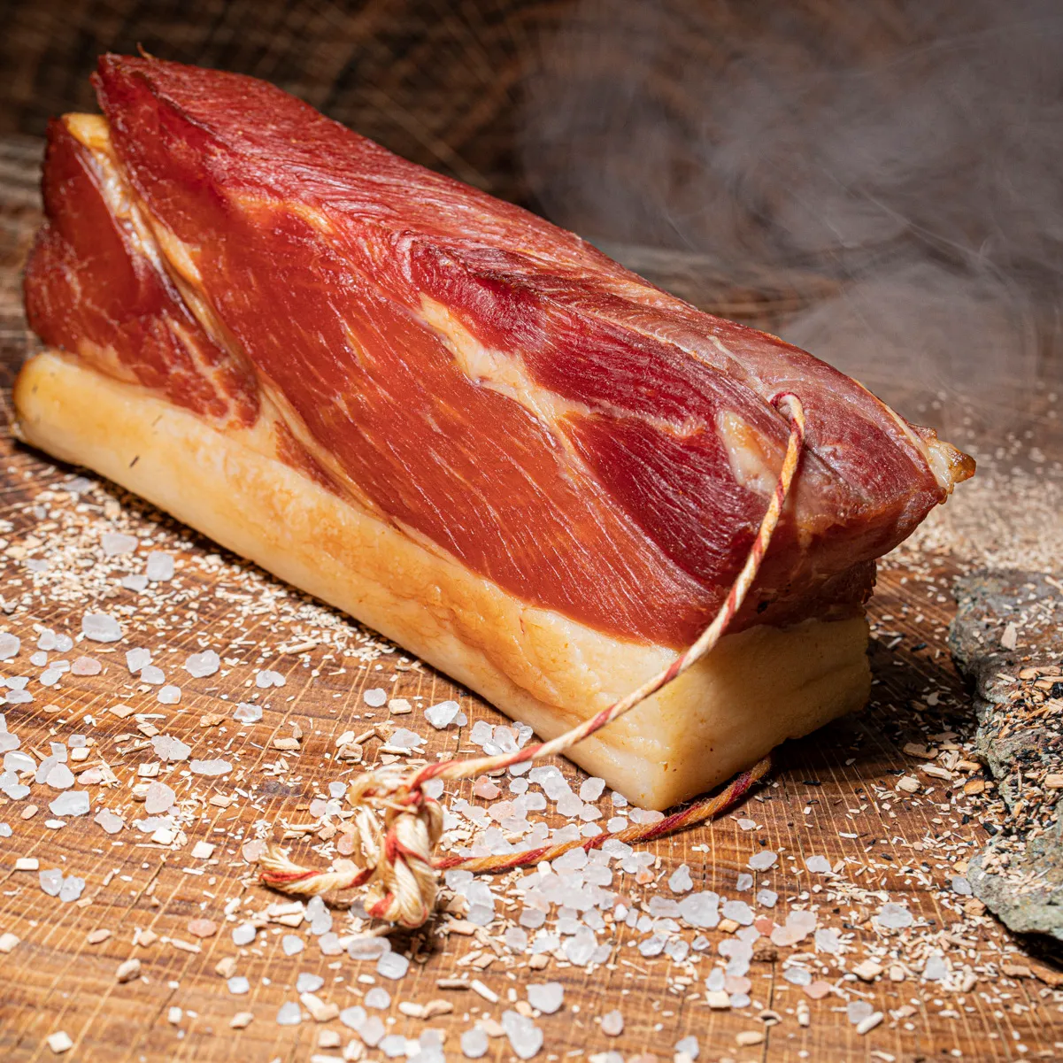 Geräucherter Schinkenspeck auf Holztisch mit Buchenspähnen, Salz und Rauch im Hintergrund.