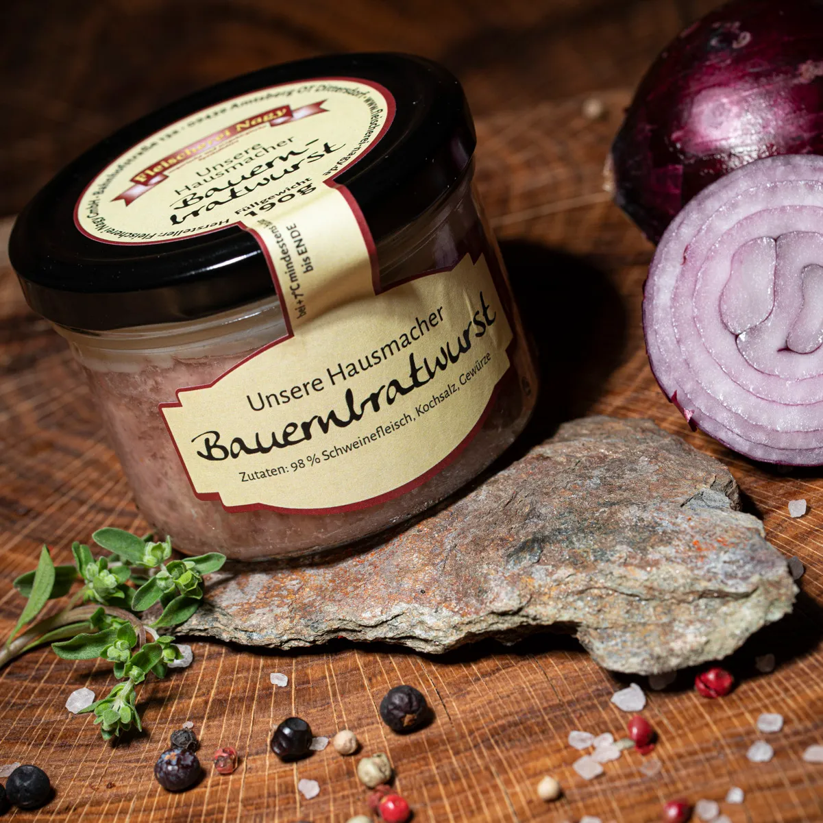 Hausmacher Bauernbratwurst im Glas auf einem Holzbrett mit halbierter roter Zwiebel als Deko.