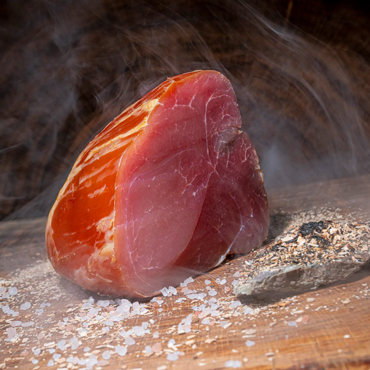 Unsere Delikatesse, feinen geräucherten Nussschinken kaufen  - purer Genuss!  Geräucherter halbierter Nussschinken auf Holztisch mit Buchenspähnen, Salz und Rauch im Hintergrund.