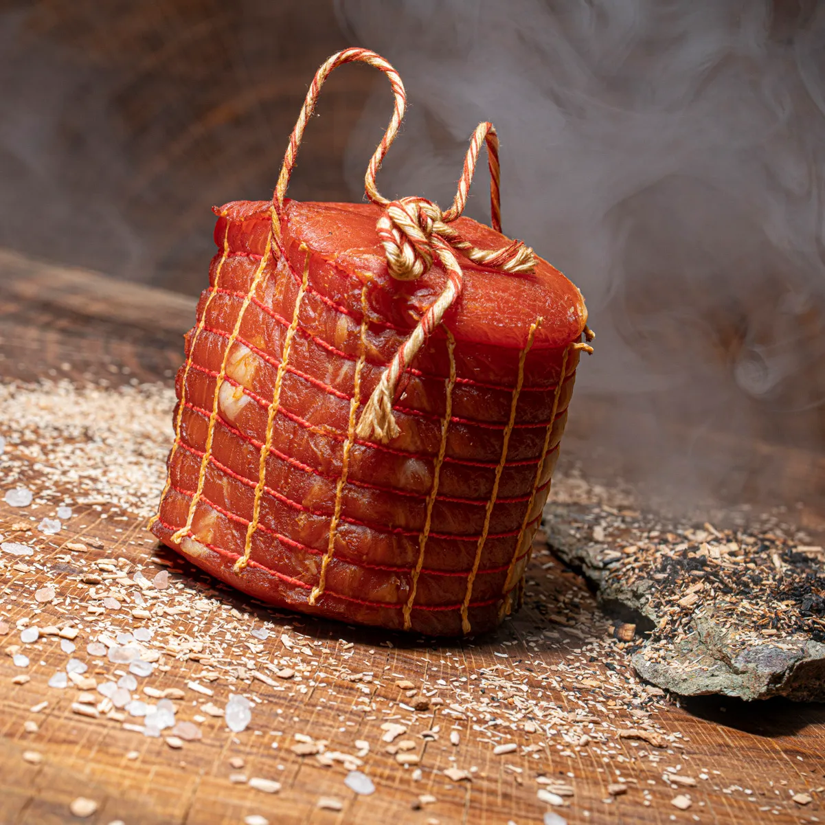 Geräucherter Rollschinken pur im Netz auf Holztisch mit Buchenspähnen und Rauch im Hintergrund.
