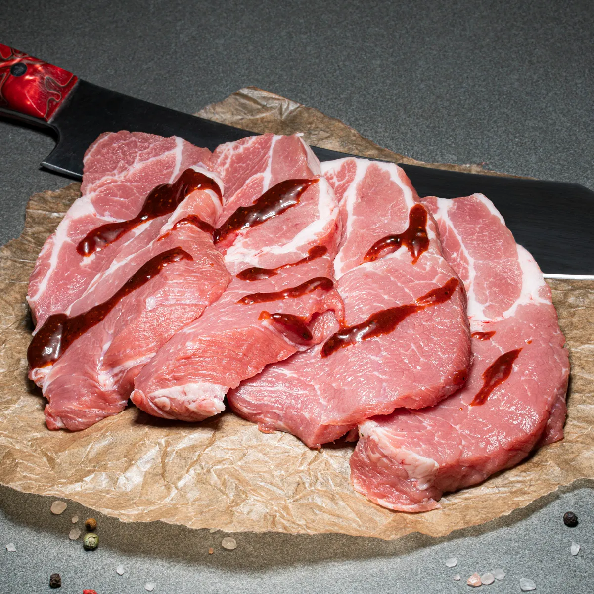 Schweinekammsteaks mit Tomaten-Pfeffer-Marinade drüber, auf Fleischpapier, im Hintergrund liegt ein Fleischmesser mit rotem Griff.