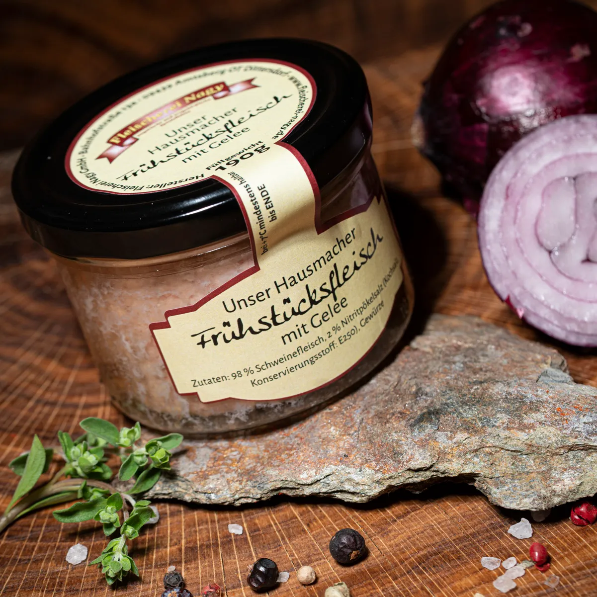 Hausmacher Frühstücksfleisch im Glas jetzt probieren und bestellen. Hausmacher Frühstücksfleisch im Glas auf einem Holzbrett mit halbierter roter Zwiebel als Deko.