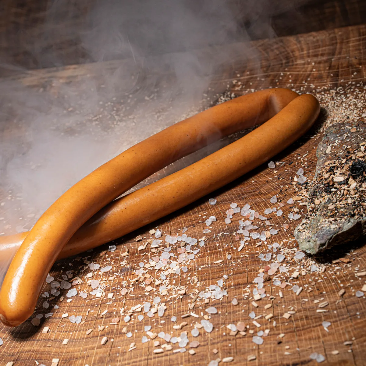Unser idealer Snack für jeden Anlass - die Wiener Würstchen! Ein Paar Wiener mit Rauch im Hintergrund auf einem Holztisch mit Salz und Holzspähnendeko.