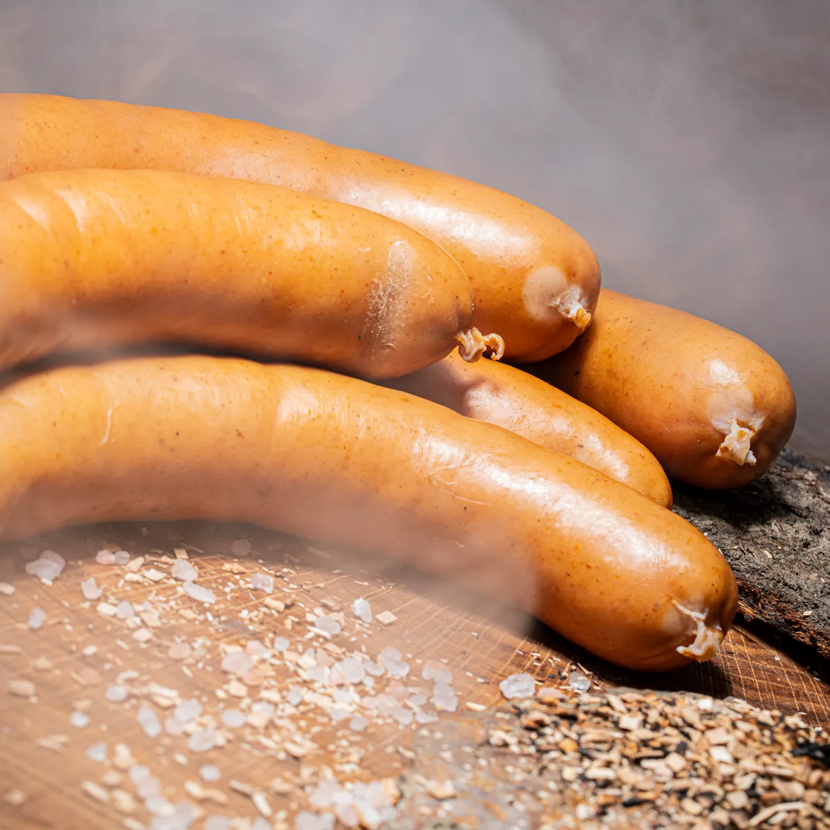 Knackige Bockwurst fein geräuchert ist der ideale Snack! 5 Stück Bockwurst mit Rauch im Hintergrund auf einem Holztisch mit Salz und Holzspähnendeko.