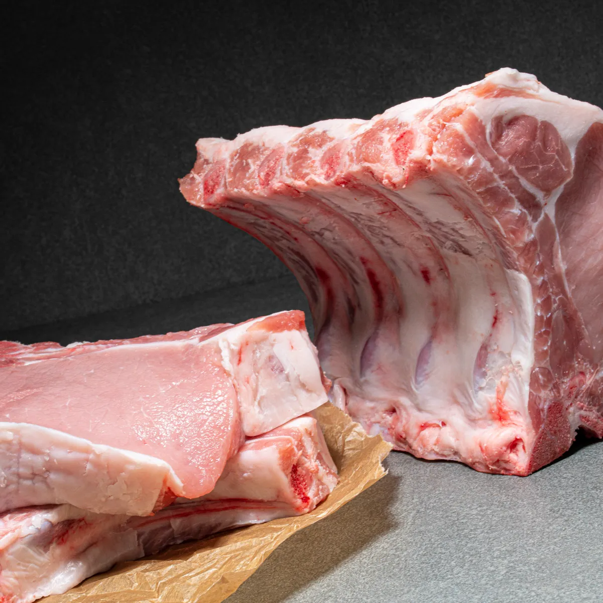 Kotelett vom Schwein am Knochen, zwei aufgeschnittene Koteletts liegen davor auf Fleischpapier.